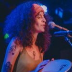 Mulher jovem, com faixa rosa, nos cabelos médios catanhos encaracolados, tocando um pandeiro em frente a um microfone.